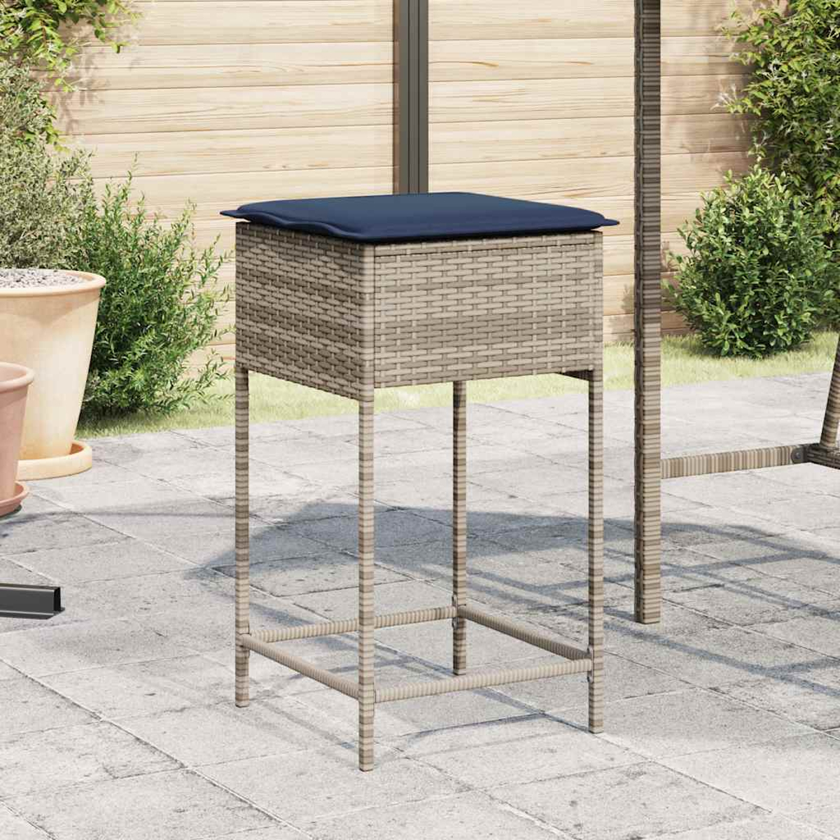 Ein Barhocker aus Korbgeflecht mit blauem Kissen auf einer Terrasse. Im Hintergrund ein Holzzaun, Topfpflanzen und Grün.