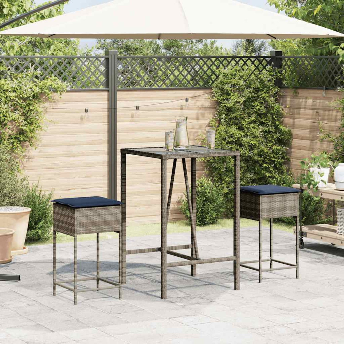 Ein Terrassenset mit Tisch, zwei Hockern und einem Sonnenschirm. Auf dem Tisch stehen ein Krug und Gläser. Der Hintergrund ist ein Holzzaun.