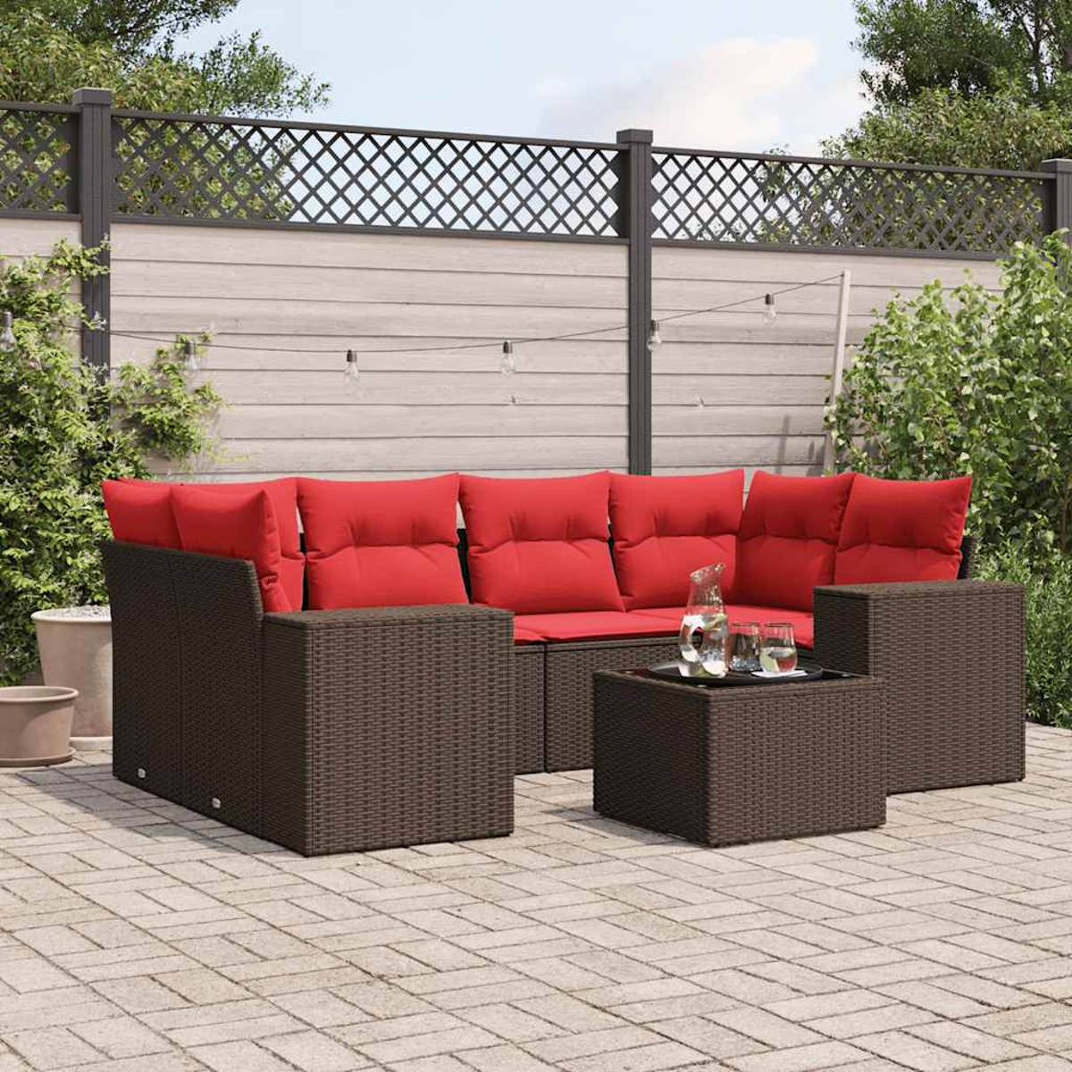 Gartenmöbel aus Korbgeflecht mit roten Kissen. Ein Tisch mit Gläsern und einer Kanne. Hintergrund: Zaun, Pflanzen und Lichterketten.