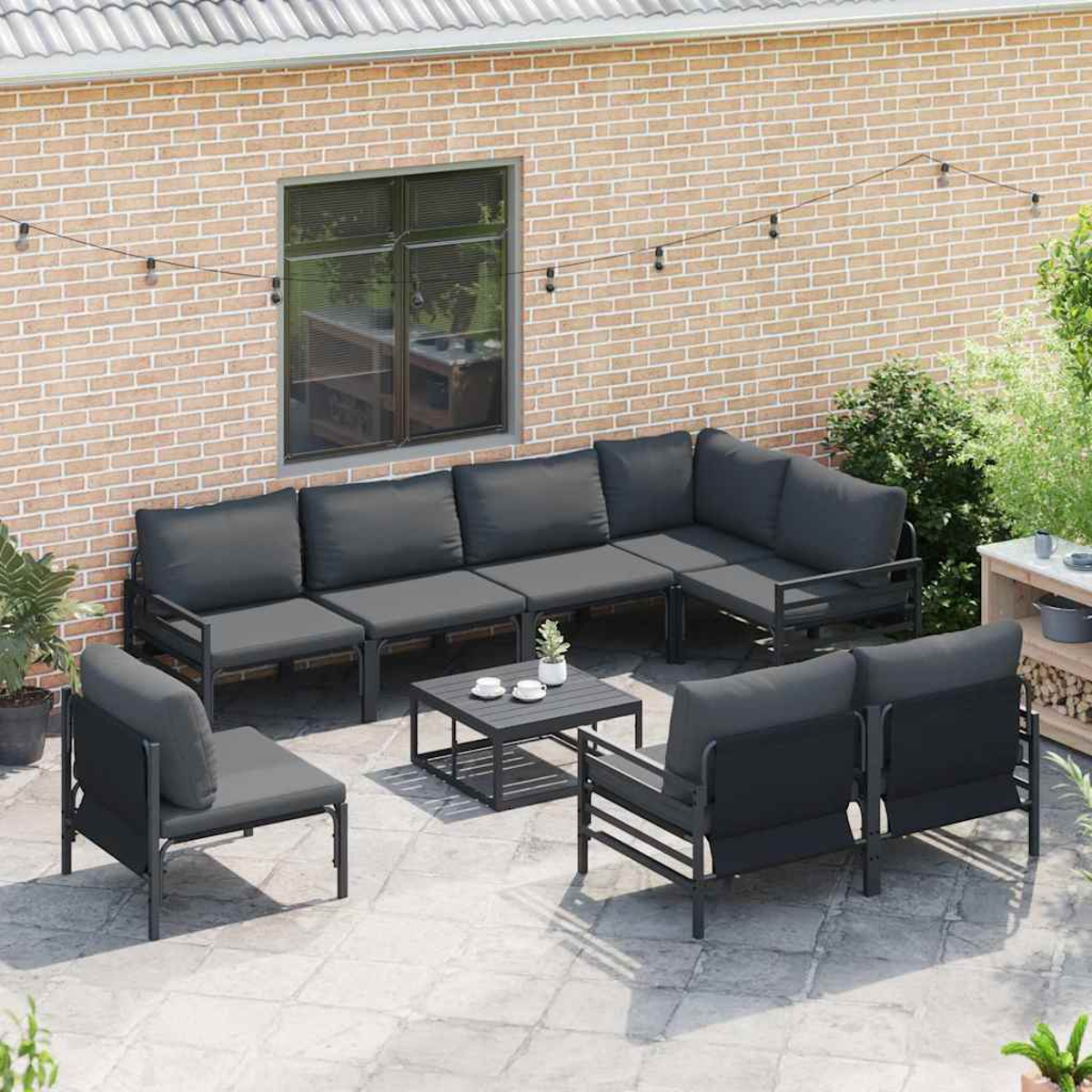 Außenbereich mit dunkelgrauem Ecksofa, Stühlen und einem Couchtisch auf einer Steinterrasse. Ziegelmauer im Hintergrund.