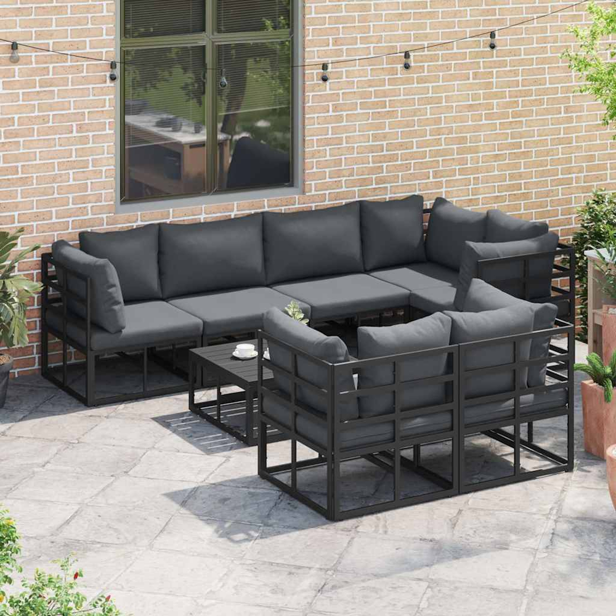 Garten-Sitzgruppe mit dunkelgrauen Kissen und schwarzen Rahmen auf einer Terrasse, neben einer Backsteinmauer, mit einem Couchtisch.