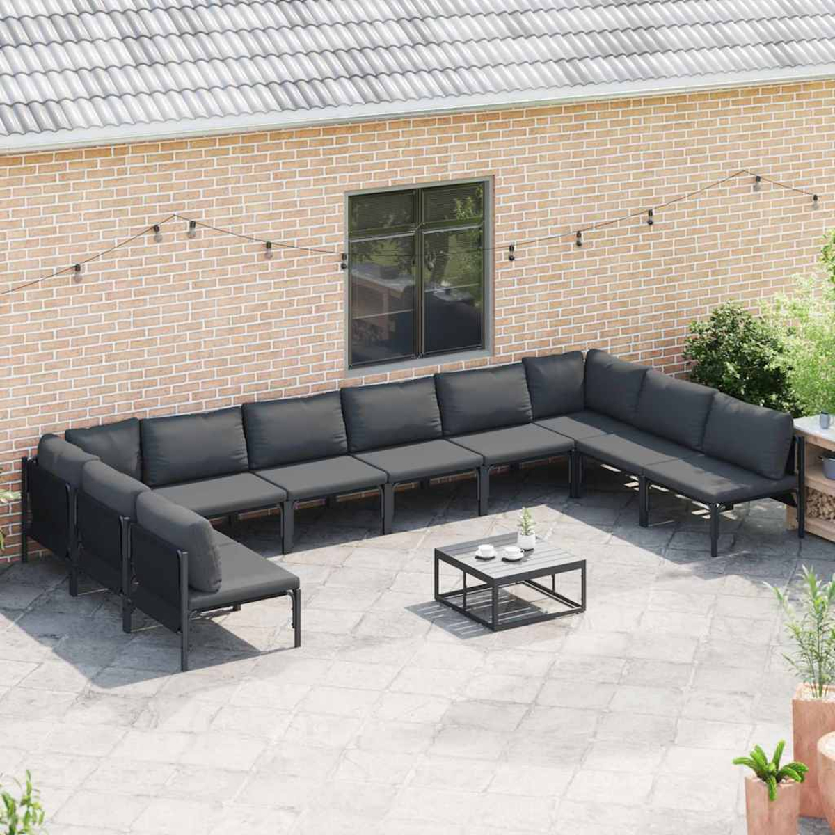 Un patio esterno con un grande divano grigio a forma di U, un tavolino con tazze e un muro di mattoni sullo sfondo.