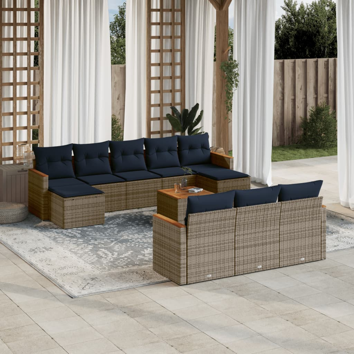 Außenbereich mit einem geflochtenen Sofa, blauen Kissen und einem Holztisch, auf einer Terrasse mit Vorhängen und Grün im Hintergrund.