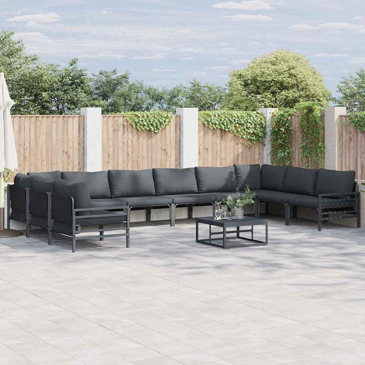 Set patio esterno con cuscini grigi e tavolo su un patio piastrellato. Recinzione in legno e piante verdi sullo sfondo.