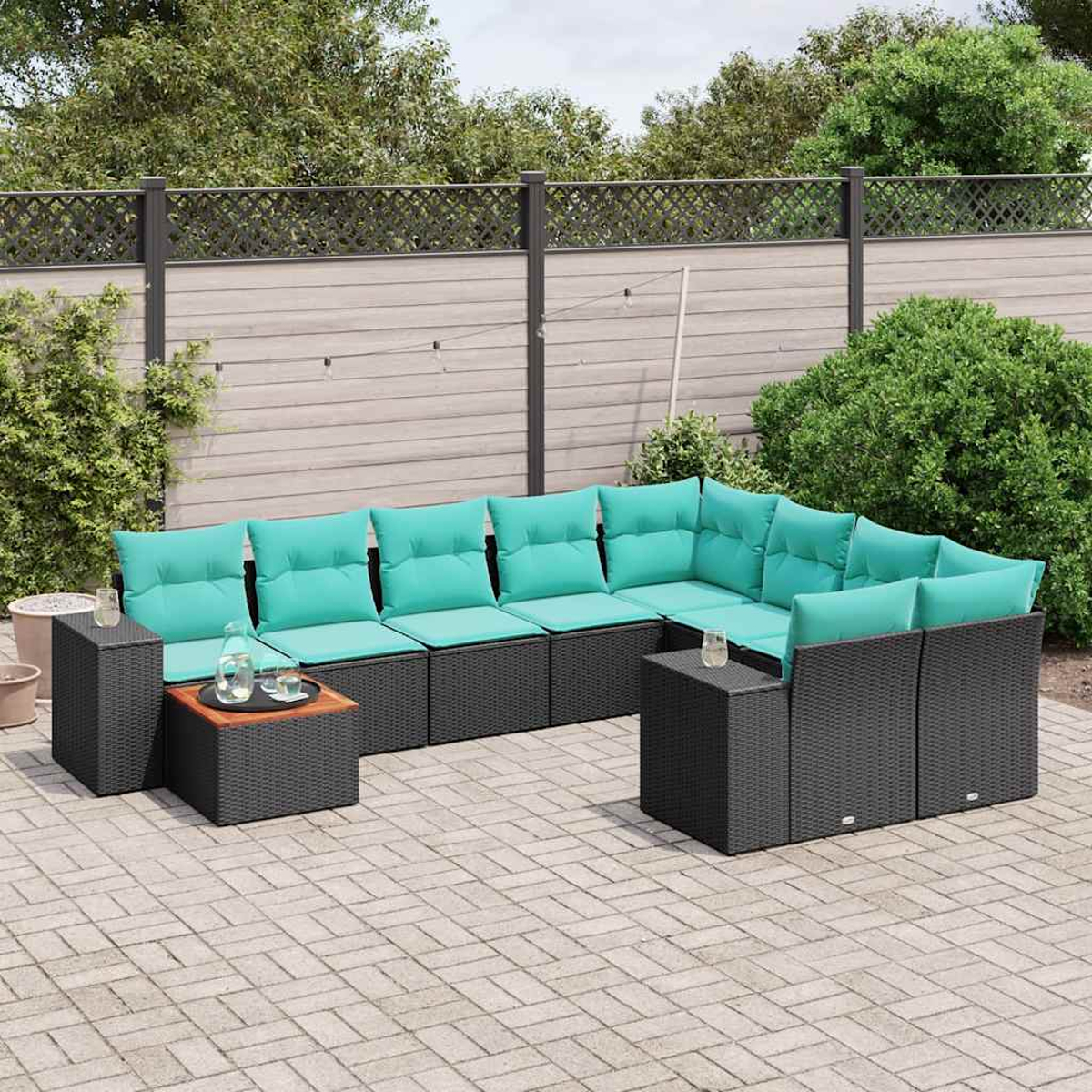 Ein U-förmiges Outdoor-Sofa mit tealfarbenen Kissen, schwarzen Korbflechtrahmen und kleinen Tischen. Auf einer Ziegelterrasse mit Zaun und Grün.