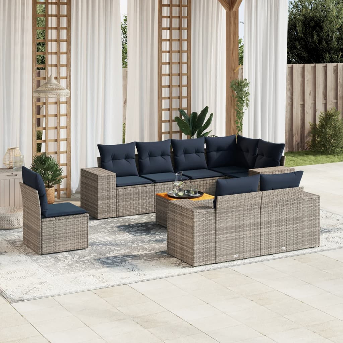 Outdoor-Sitzgruppe: graue Korbmöbel mit marineblauen Kissen auf einem Teppich. Weiße Vorhänge, Pflanzen und Holzspaliere im Hintergrund.