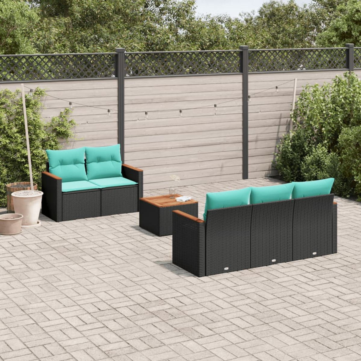 Terrassengarnitur mit schwarzer Korbmöbel, türkisfarbenen Kissen und einem Holztisch auf einer Ziegelterrasse. Grüne Pflanzen und ein Zaun sind im Hintergrund.