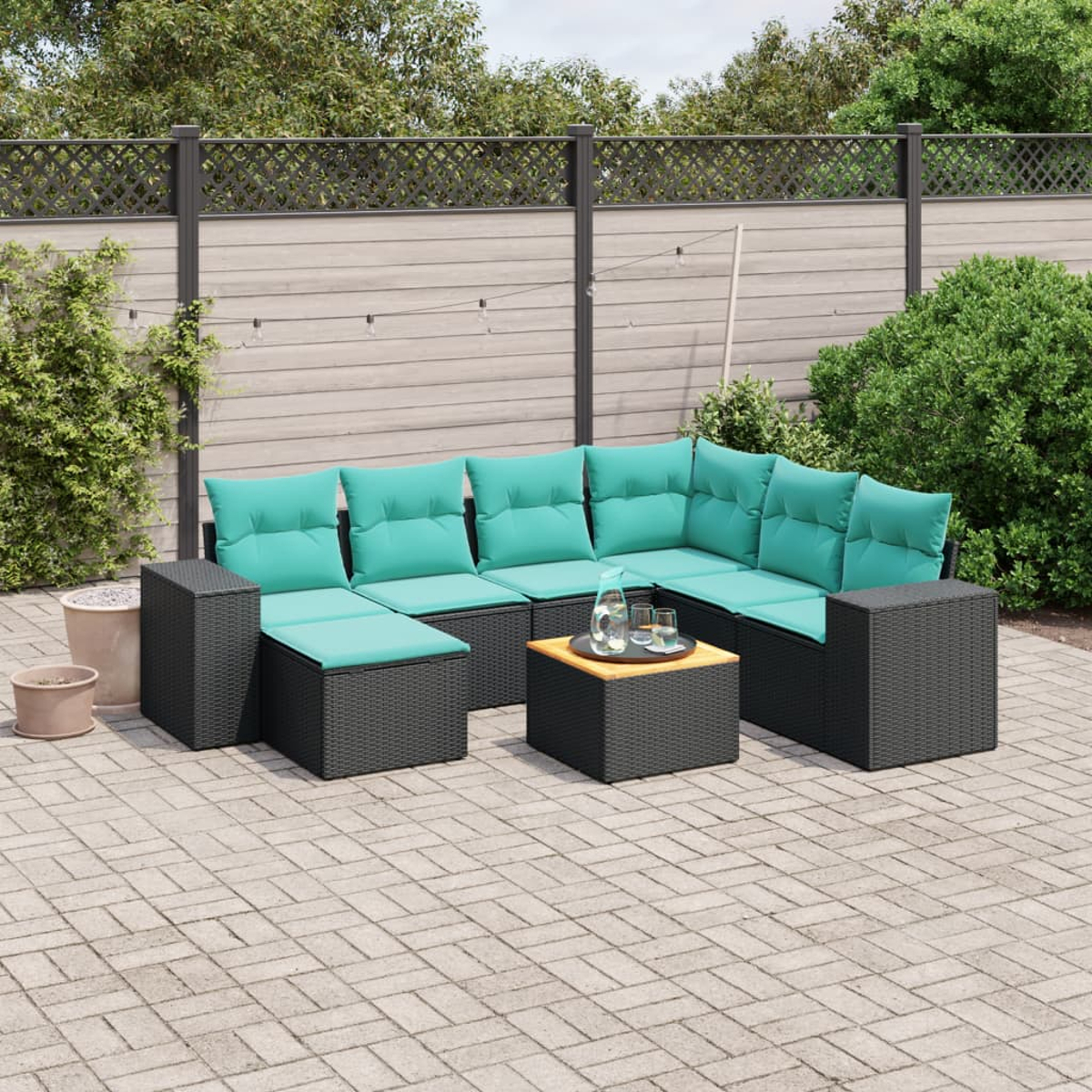 Outdoor-Ecksofa mit tealfarbenen Kissen und einem Tisch mit Gläsern auf einer Backstein-Terrasse. Im Hintergrund sind Zaun und Grün zu sehen.