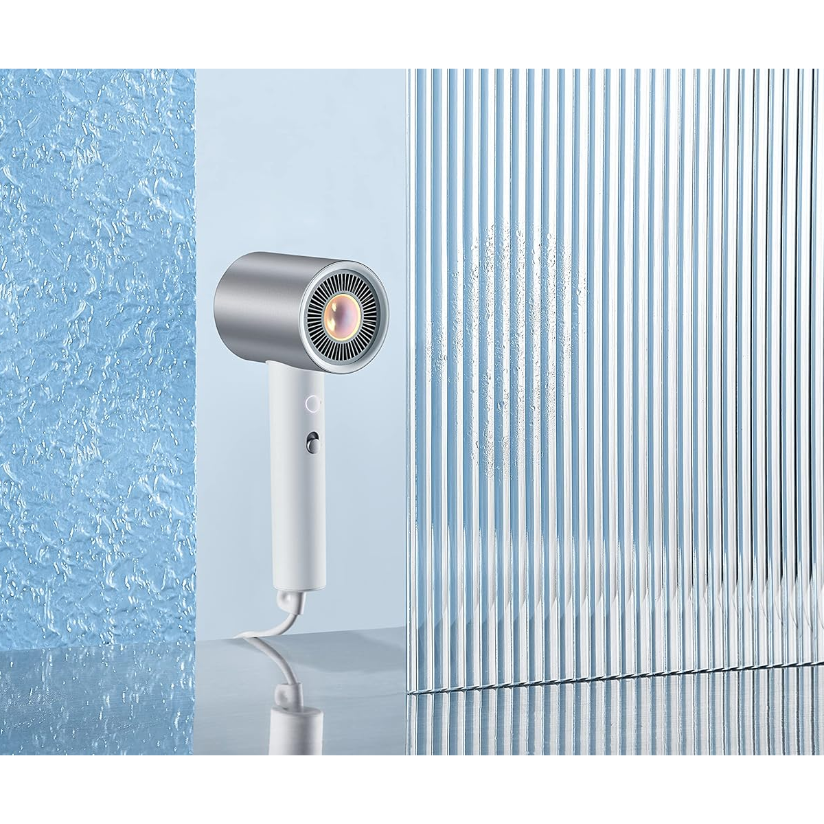 Srebrna i biała suszarka do włosów stoi na tle teksturowanej niebieskiej ściany i szklanego panelu z pionowymi paskami.