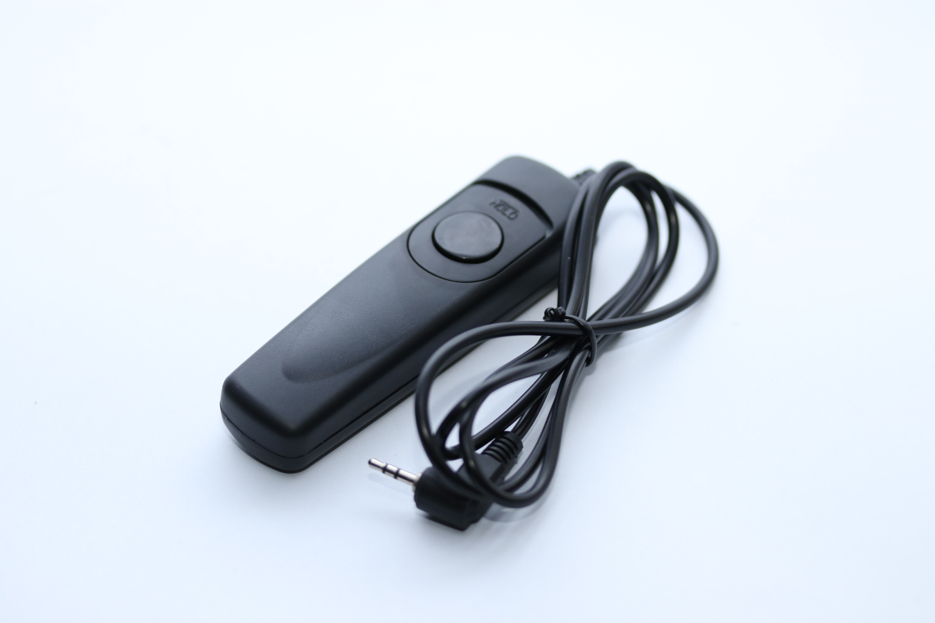 Eine schwarze Kamera-Fernbedienung mit Knopf und Kabel liegt auf einer weißen Oberfläche. Das Kabel hat einen Stecker.