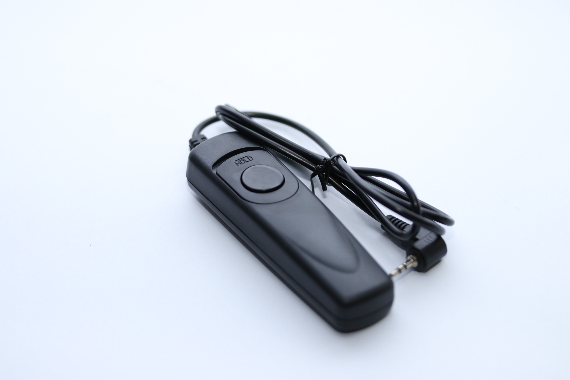 Eine schwarze Kamerafernbedienung mit Kabel und Stecker liegt auf einer weißen Oberfläche. Die Fernbedienung hat einen Knopf mit der Aufschrift 'HOLD'.