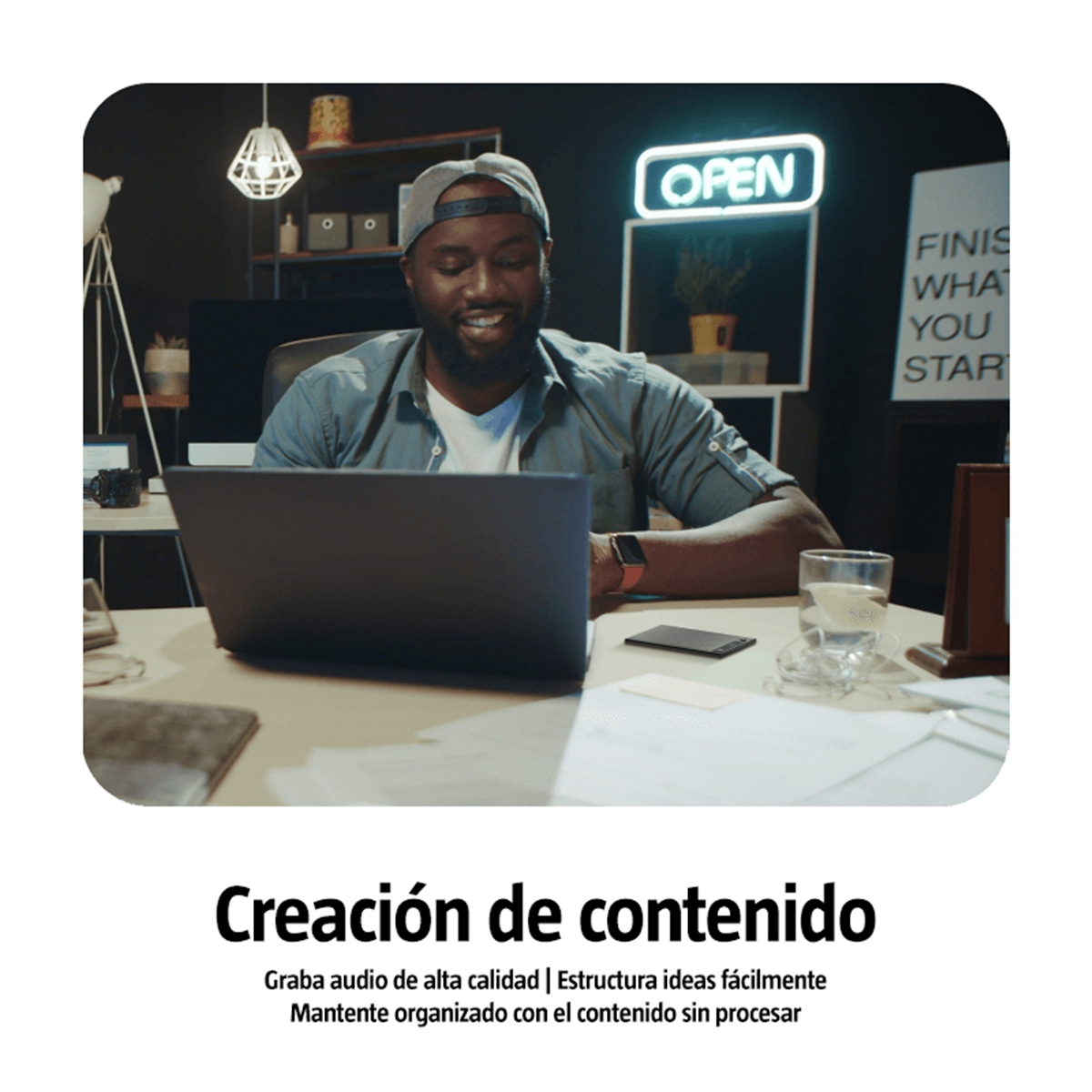 Un hombre con gorra sonríe a una laptop. Un letrero de "OPEN" está arriba. Papeles y un vaso de agua están en el escritorio.