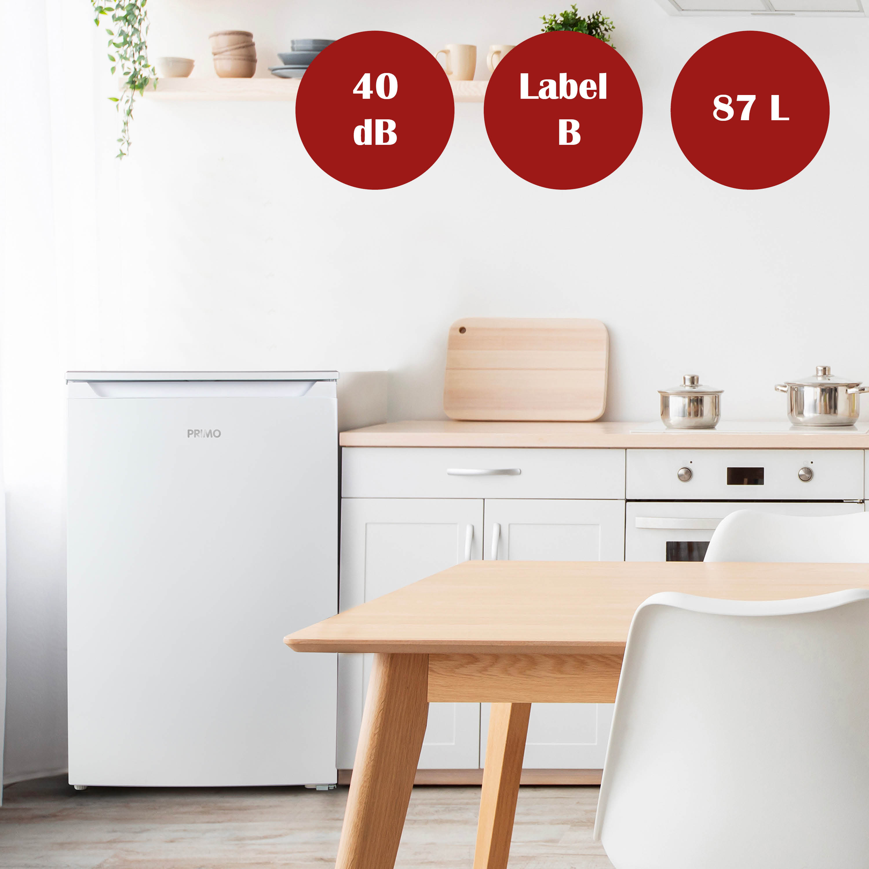 Een witte Primo-koelkast in een keuken. Labels tonen 40dB, Label B en 87L. Een tafel en stoelen ervoor.