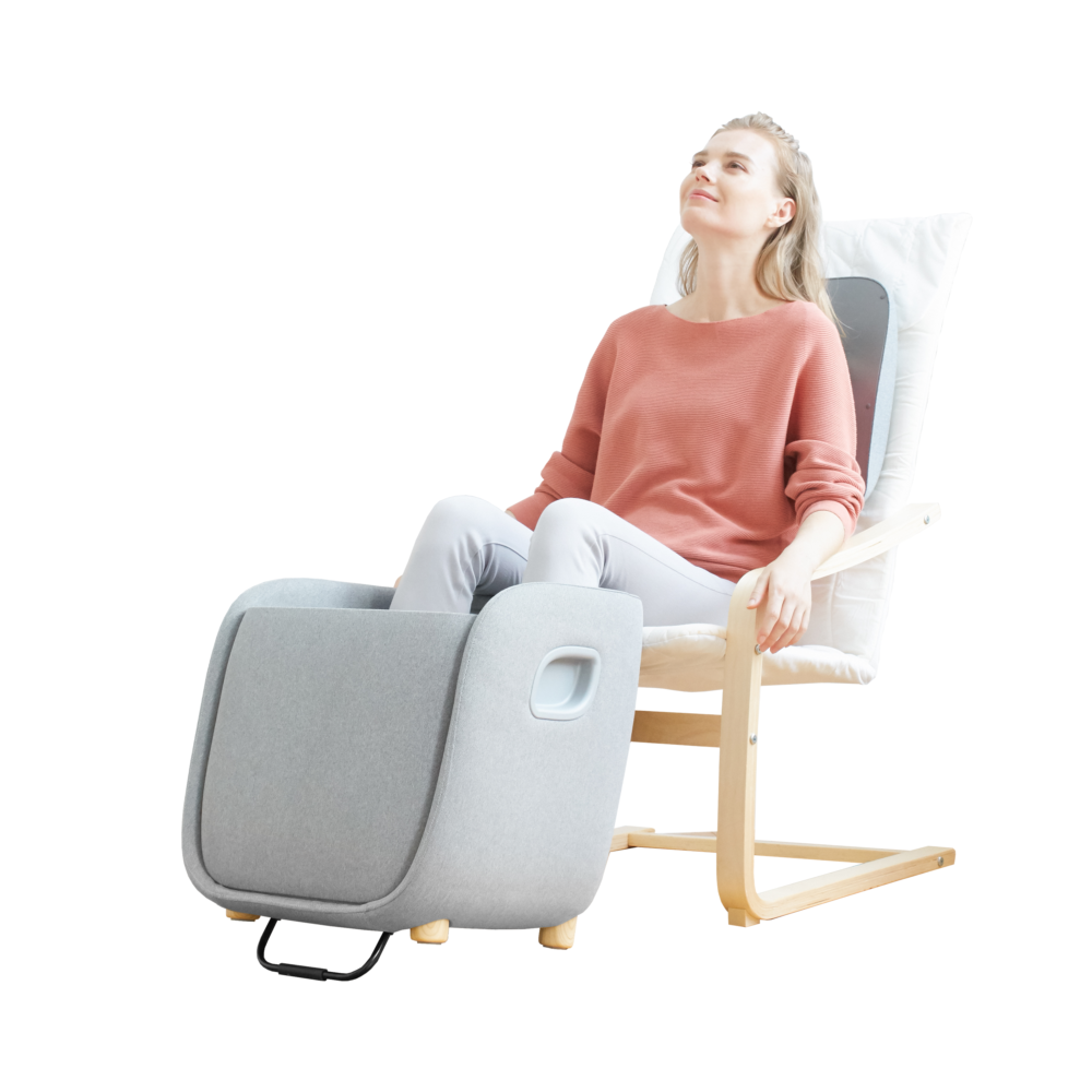 Een vrouw zit in een massagestoel en kijkt omhoog. Ze draagt een roze trui en een witte broek. De stoel is grijs met houten poten.