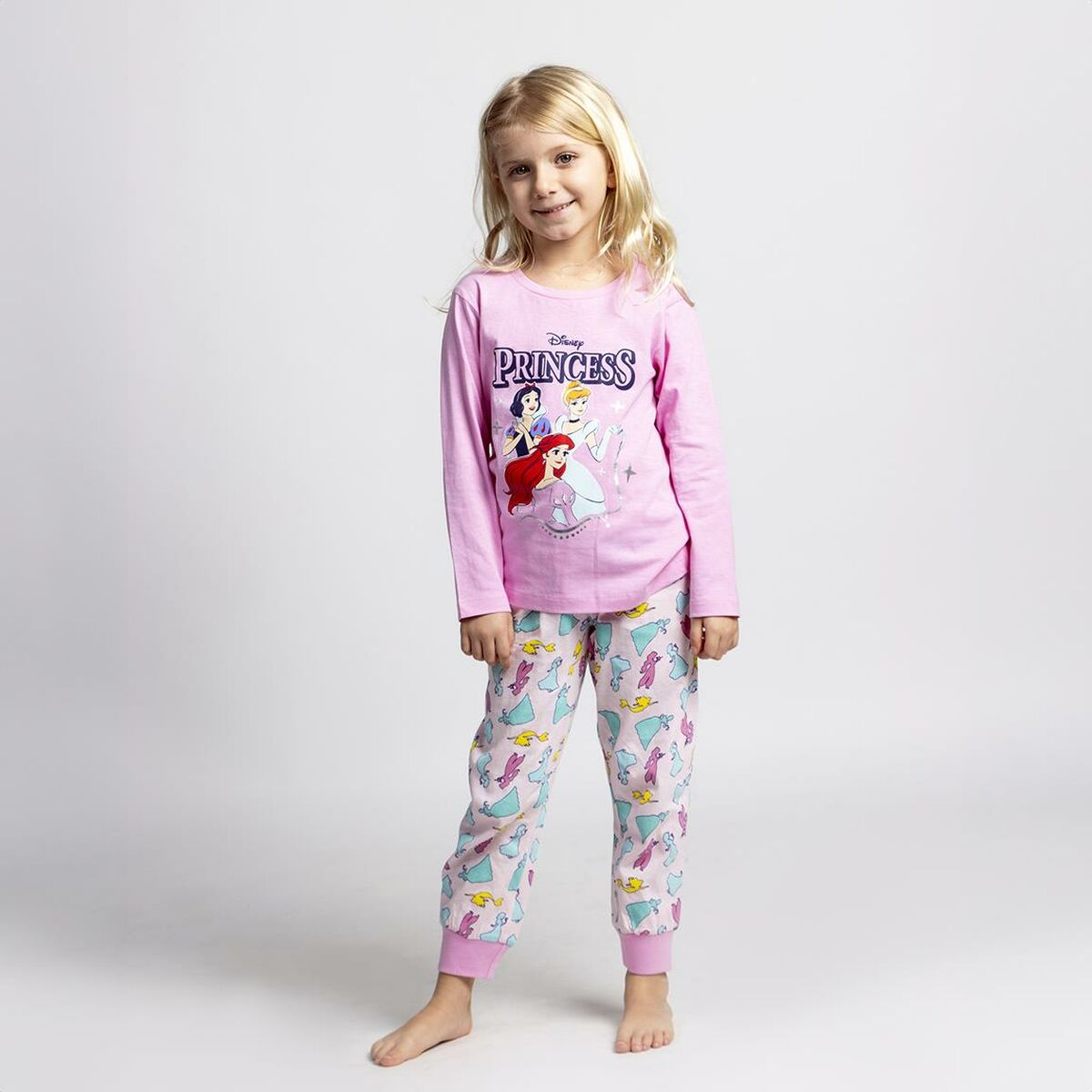 Ein junges Mädchen in rosa Disney Prinzessinnen-Pyjamas steht und lächelt. Das Hemd hat Prinzessinnen-Grafiken. Der Hintergrund ist schlicht weiß.