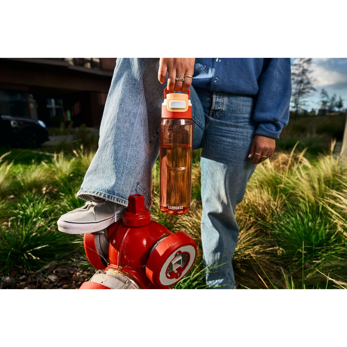 Person hält eine Wasserflasche und steht in der Nähe eines roten Hydranten. Sie trägt Jeans und einen blauen Pullover, im Hintergrund Gras.