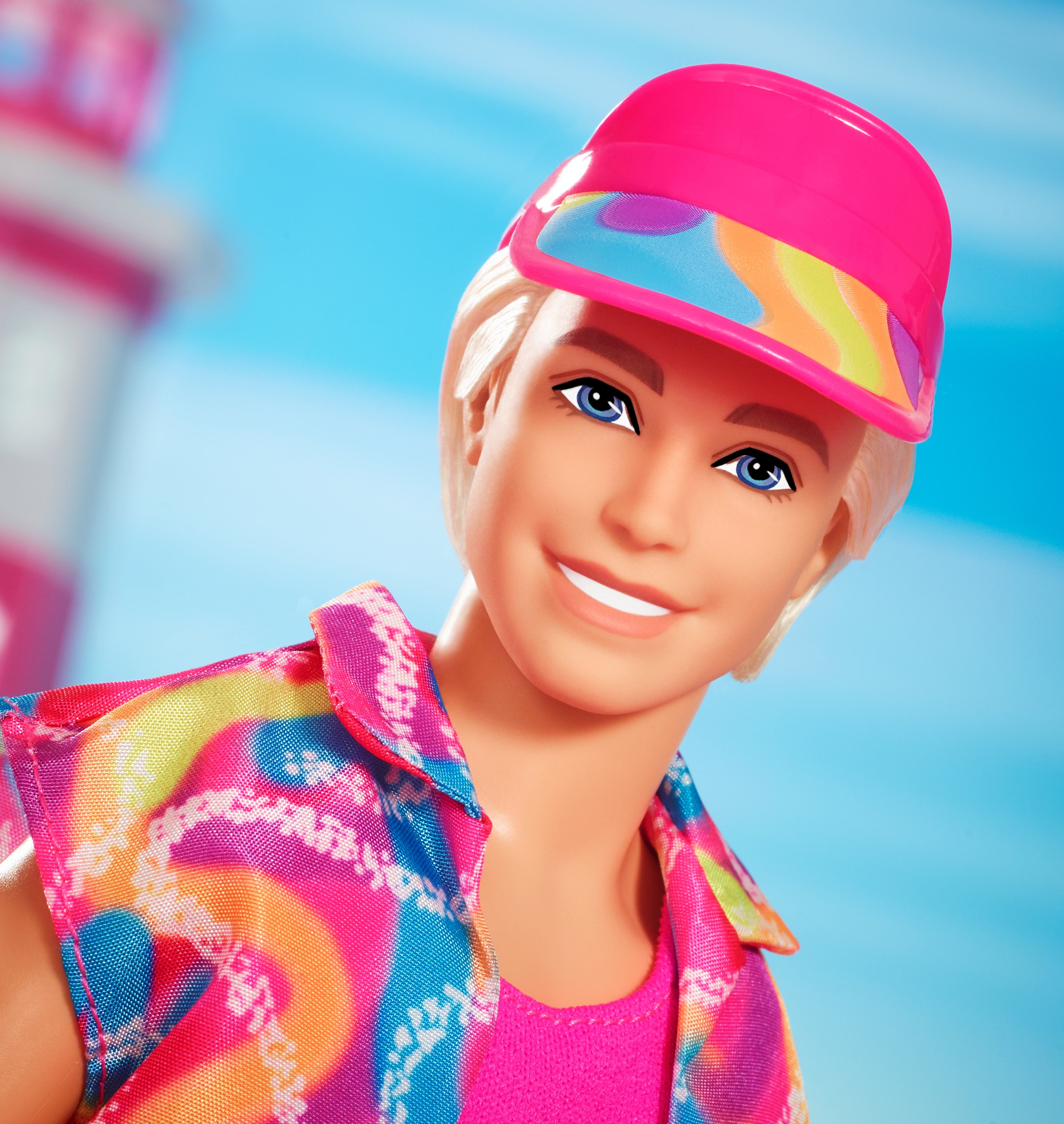 Muñeco Ken con cabello rubio, visera rosa y camisa con diseños de arcoíris. Cielo azul al fondo.