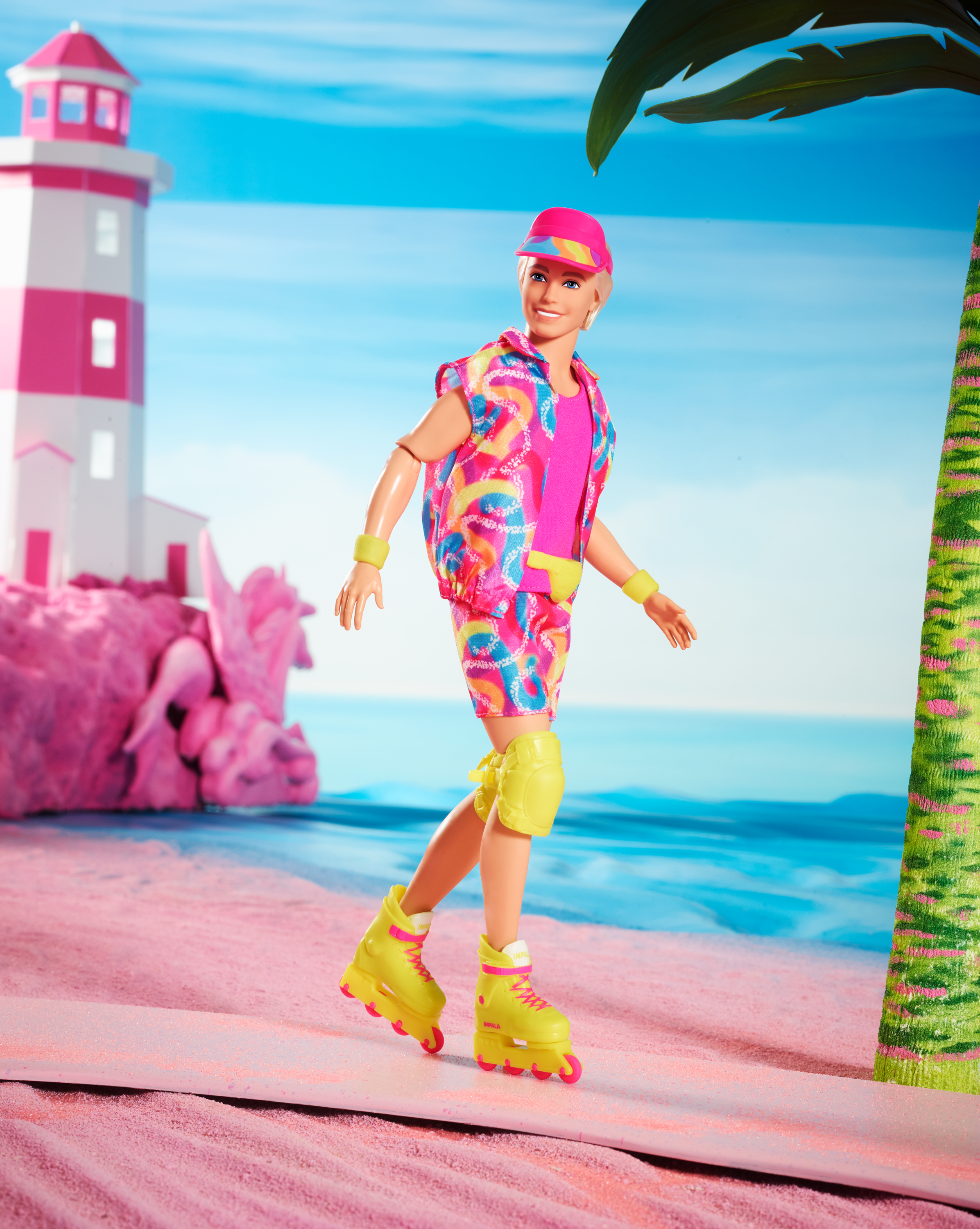 Muñeco Ken en patines, con visera rosa, de pie en una playa rosa. Hay un faro y una palmera en el fondo.