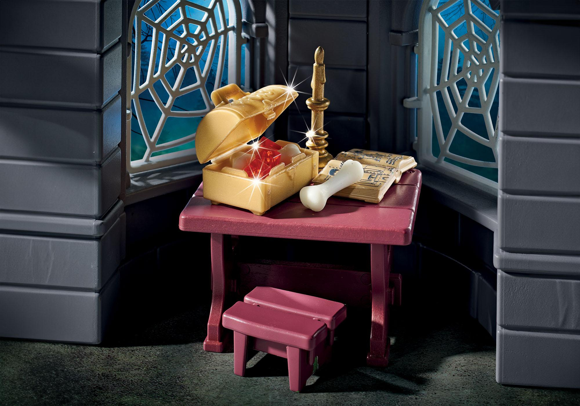 Scena ze skrzynią skarbów, kością, świecą i książką na stole, obok okna z pajęczyną.