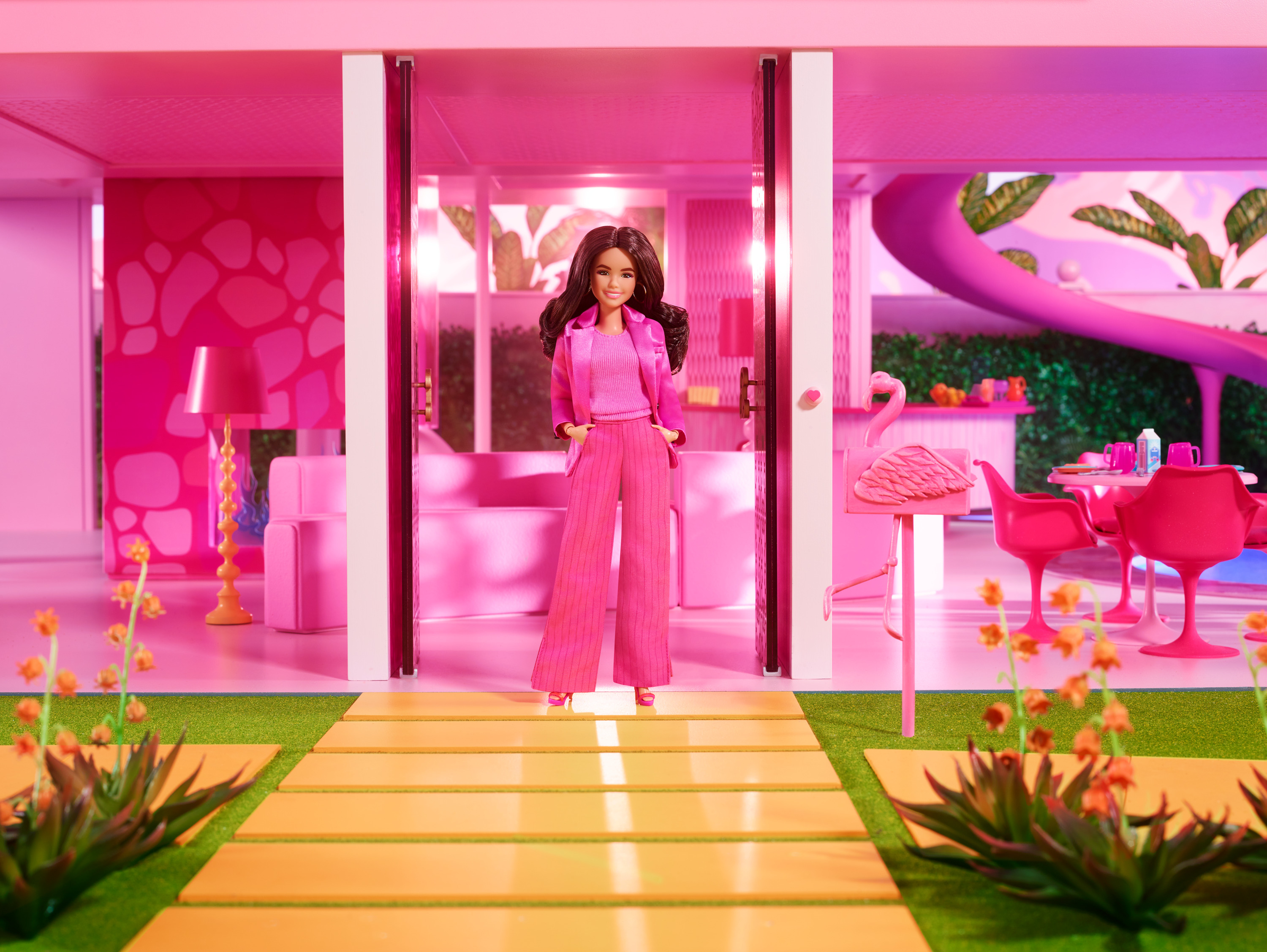 Eine Barbie-Puppe steht in einem rosa Haustür. Sie hat ein rosa Outfit an. Eine Flamingostatue und ein Pool sind sichtbar.