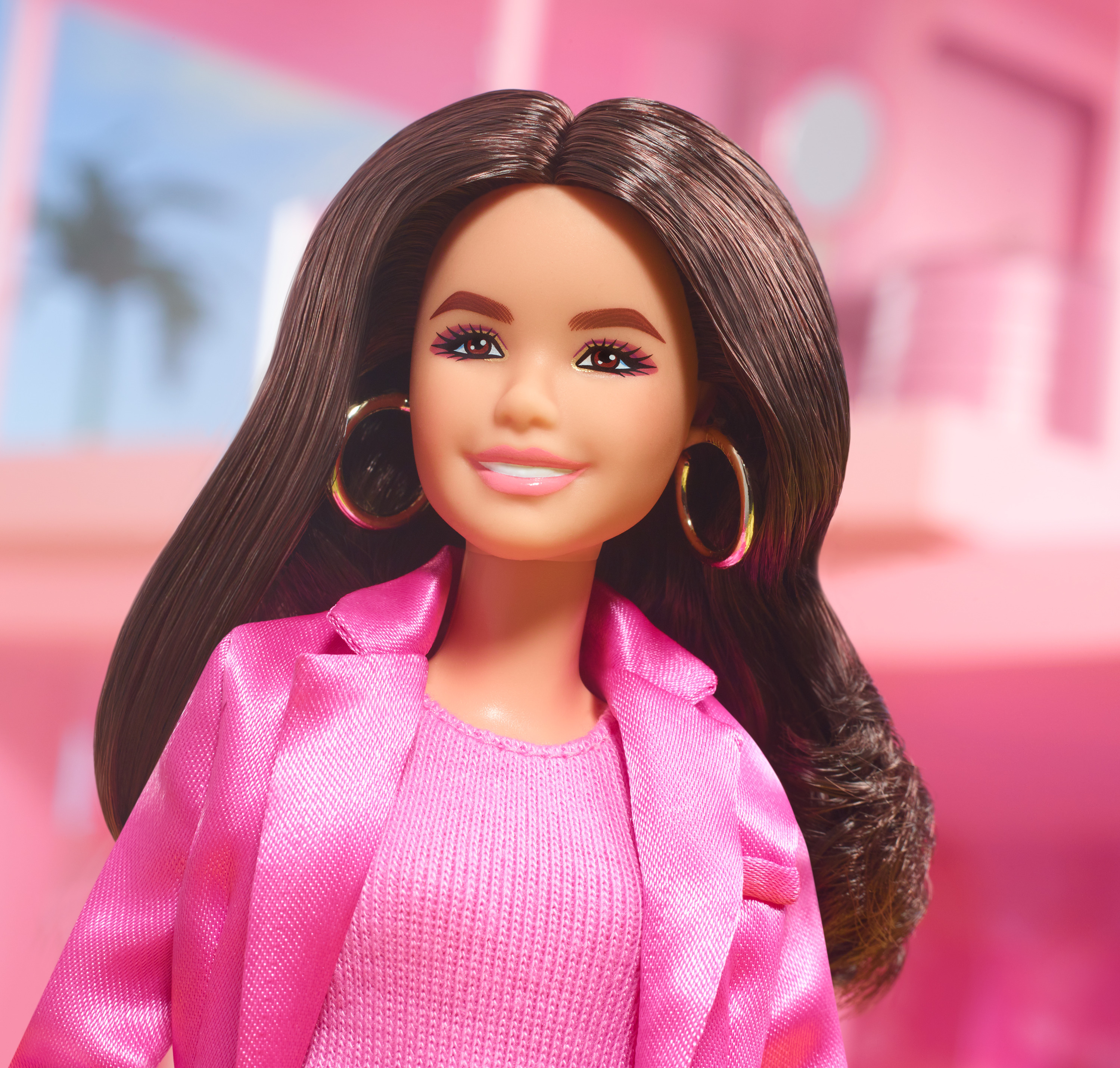 Eine Barbie-Puppe mit braunem Haar, goldenen Ohrringen und einem rosa Anzug. Der Hintergrund ist ein verschwommener rosafarbener Raum.