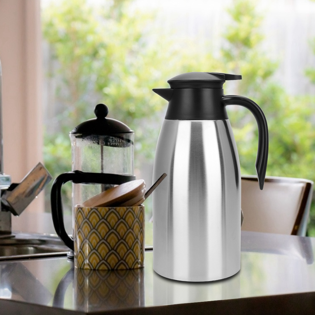 Eine Edelstahl-Thermoskanne und eine Kaffeepresse stehen auf einem Tisch. Eine gemusterte Tasse und grünes Laub sind im Hintergrund.