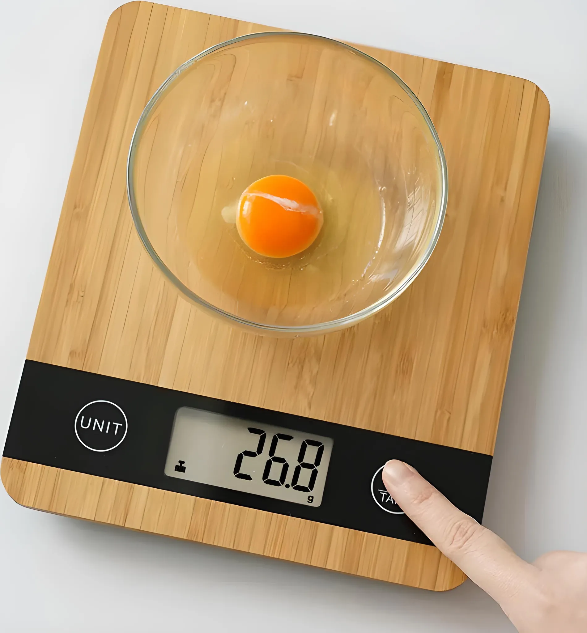 Eigelb in einer Glasschüssel auf einer Waage. Die Waage zeigt 26,8 g an. Ein Finger drückt die Tara-Taste. Holzoberfläche.