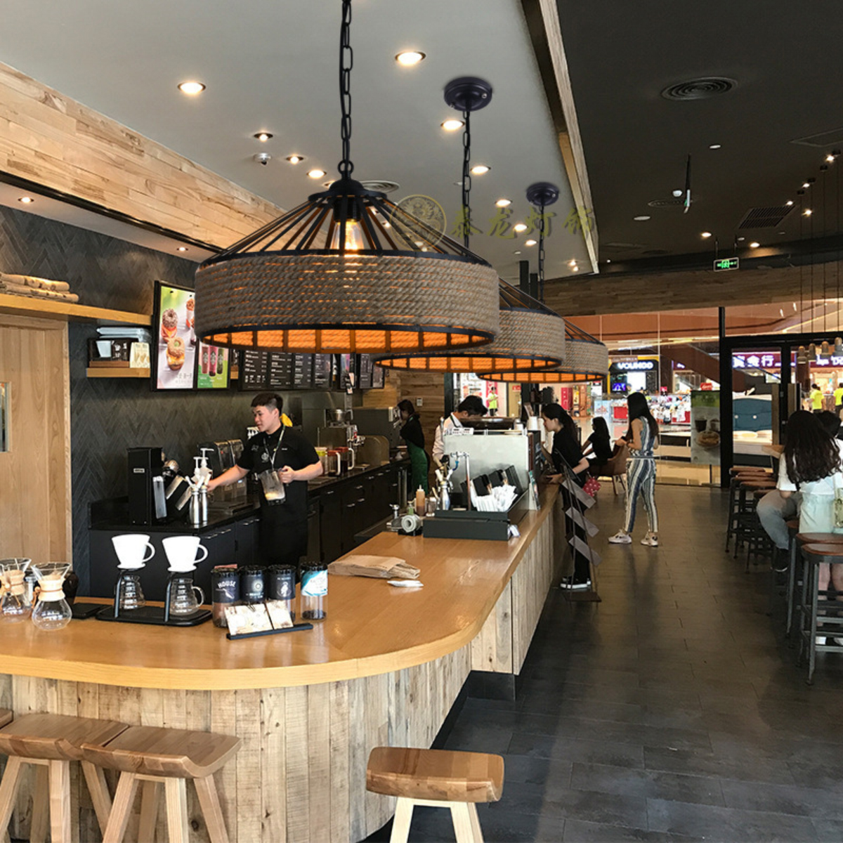 Ein Café mit Holzkühltheke, Hockern und Hängelampen. Leute bestellen und arbeiten hinter der Theke. Ein Einkaufszentrum ist sichtbar.