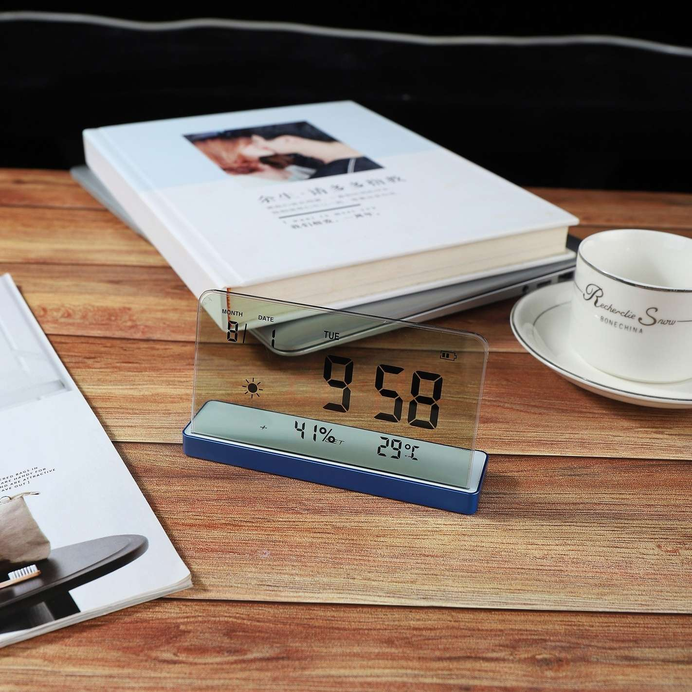 Eine Digitaluhr zeigt 9:58 Uhr an. Daneben befinden sich eine Teetasse, Bücher und eine Zeitschrift auf einer Holzoberfläche. Die Uhr zeigt Wetterdetails.