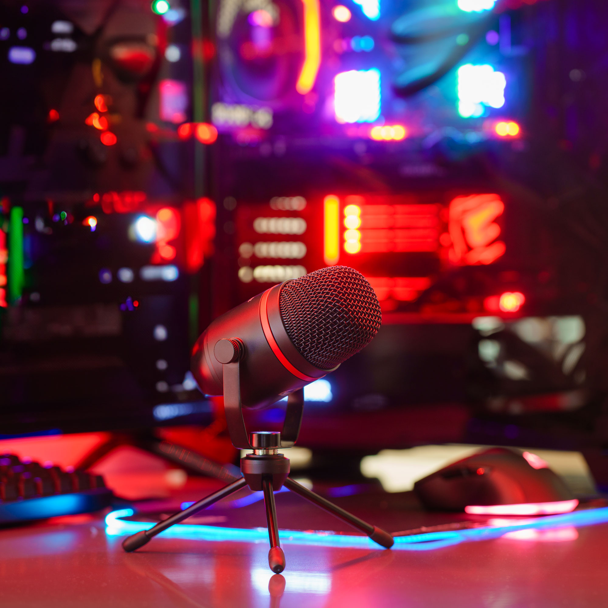 Czarny mikrofon na statywie przed komputerem z czerwonymi i niebieskimi światłami oraz klawiaturą.