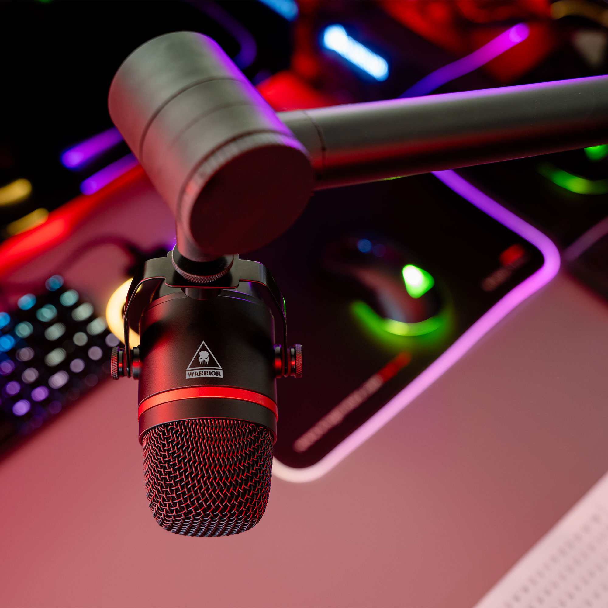 Czarny mikrofon z czerwonym akcentem zawieszony nad podświetlaną klawiaturą i podkładką pod mysz. Scena skąpana w kolorowych światłach.