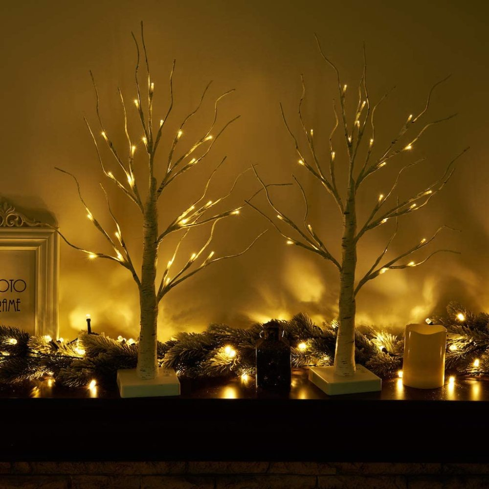Zwei beleuchtete dekorative Bäume mit Lichterketten. Eine Kerze und ein Rahmen sind ebenfalls auf dem Mantel zu sehen, mit einer Girlande.