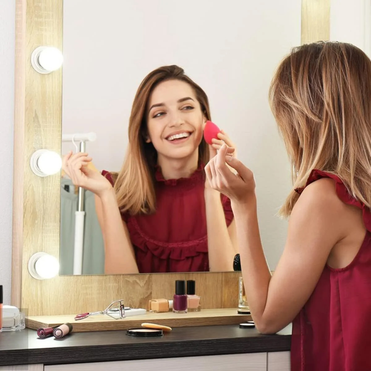 Frau, die sich vor einem Spiegel mit einem Schwamm schminkt. Schminktisch mit Make-up-Produkten. Rotes Kleid und glücklicher Gesichtsausdruck.