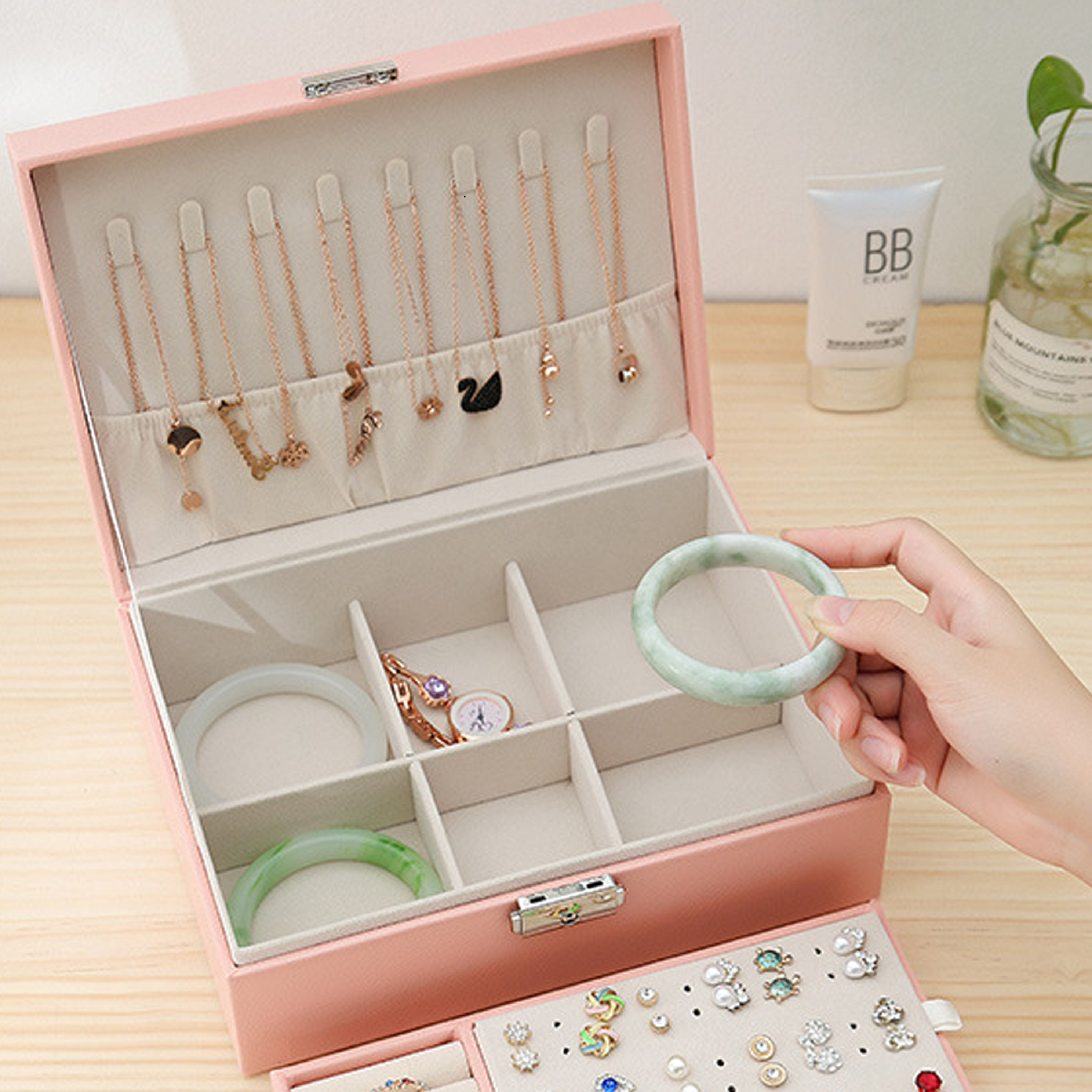 Offene rosa Schmuckschatulle mit Halsketten, Armbändern und Ohrringen. Eine Hand hält ein grünes Armband. BB-Creme und Pflanze im Hintergrund.