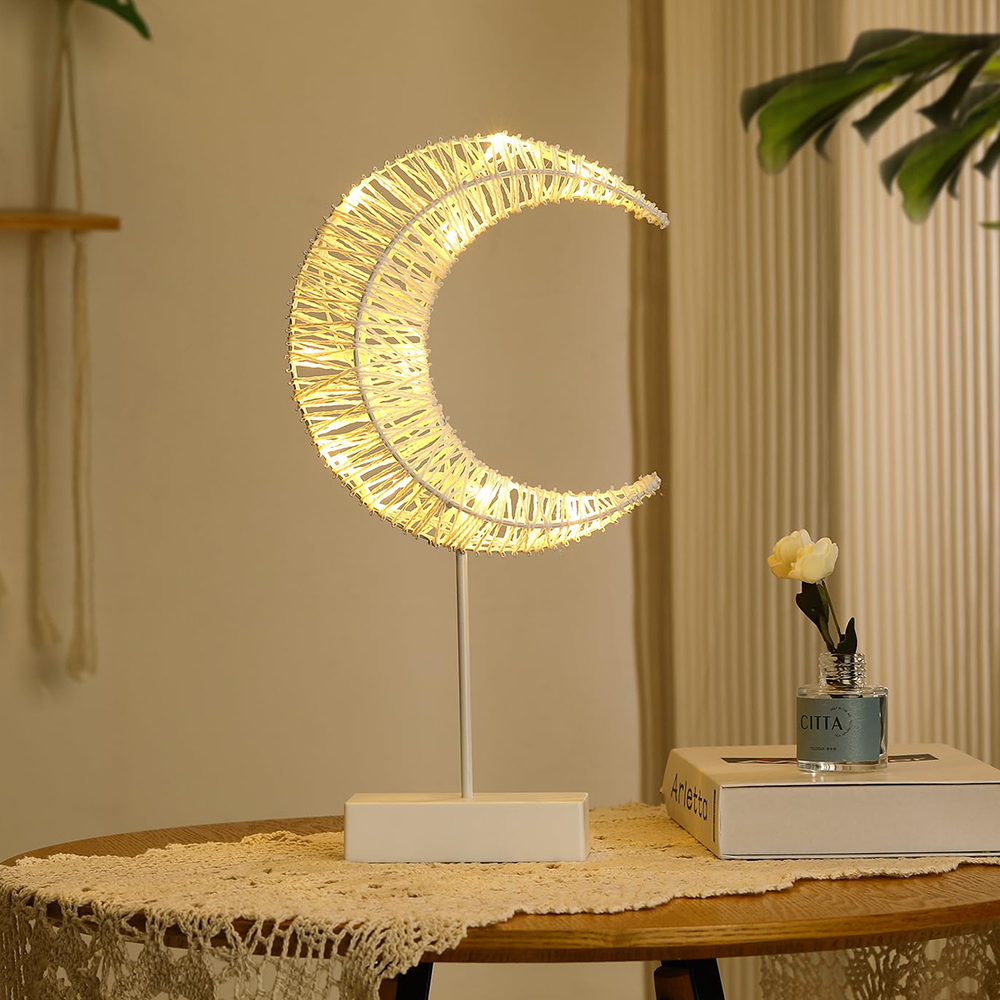 Eine halbmondförmige Lampe steht auf einem Tisch. Eine Vase mit einer Blume steht neben einem Buch. Der Tisch ist mit einem Ziertuch bedeckt.