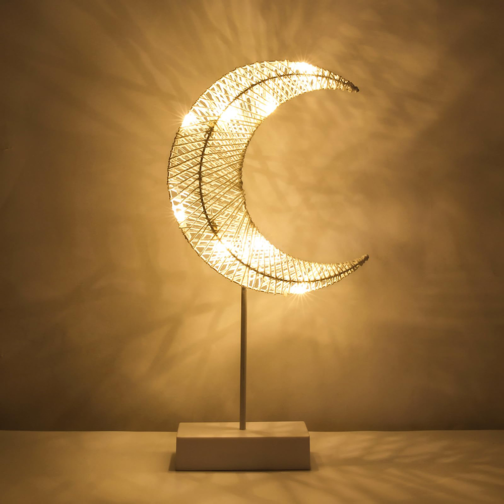 Eine Halbmondlampe mit warmen Lichtern. Sie besteht aus gewebtem Material und steht auf einem Sockel. Der Hintergrund ist eine schlichte Wand.
