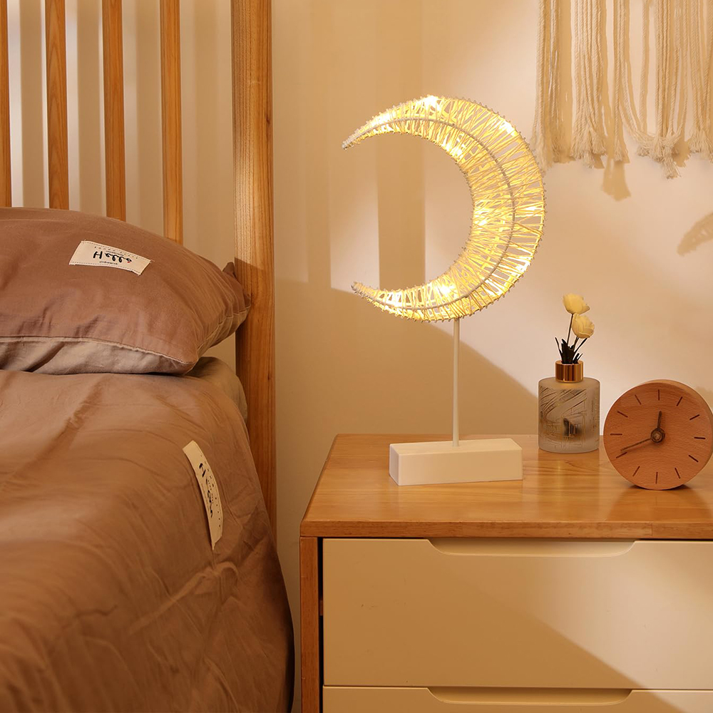 Eine Mondlampe auf einem Nachttisch. Daneben eine Vase mit Blumen und eine Uhr. Links ein Bett.