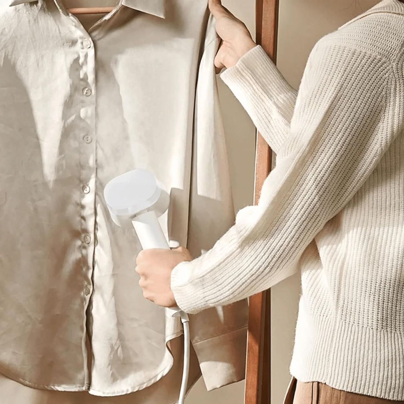 Osoba prasuje beżową koszulę białą parownicą ręczną. Osoba ma na sobie biały sweter, a koszula wisi na drewnianym wieszaku.