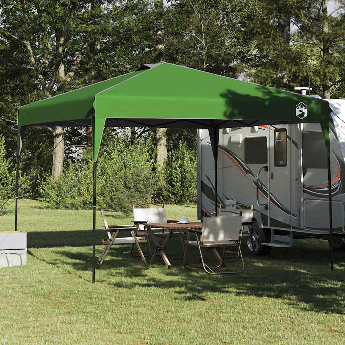 Ein grünes Vordach schützt einen Tisch und Stühle in der Nähe eines Wohnmobils. Bäume und Gras füllen den Hintergrund, was auf einen Campingplatz hindeutet.