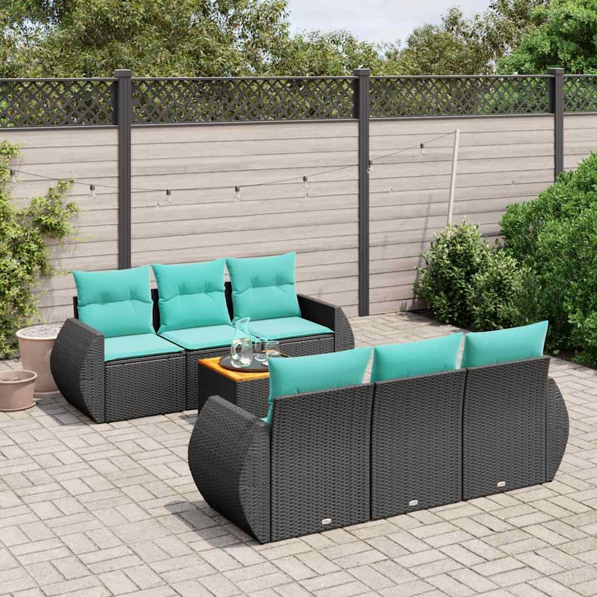 Garten-Set mit schwarzen Korbsesseln, türkisfarbenen Kissen und einem Holztisch auf einer gepflasterten Terrasse. Hintergrund ist ein Zaun und Grün.