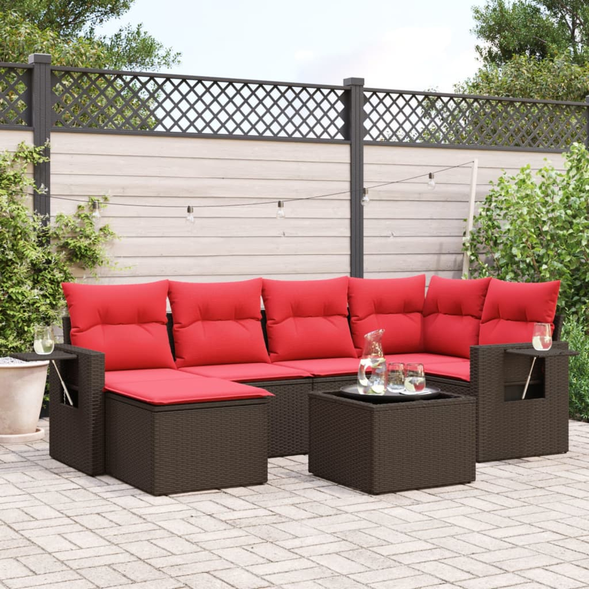 Outdoor-Ecksofa mit roten Kissen. Ein kleiner Tisch trägt Gläser und eine Kanne. Im Hintergrund ein Zaun und Pflanzen.