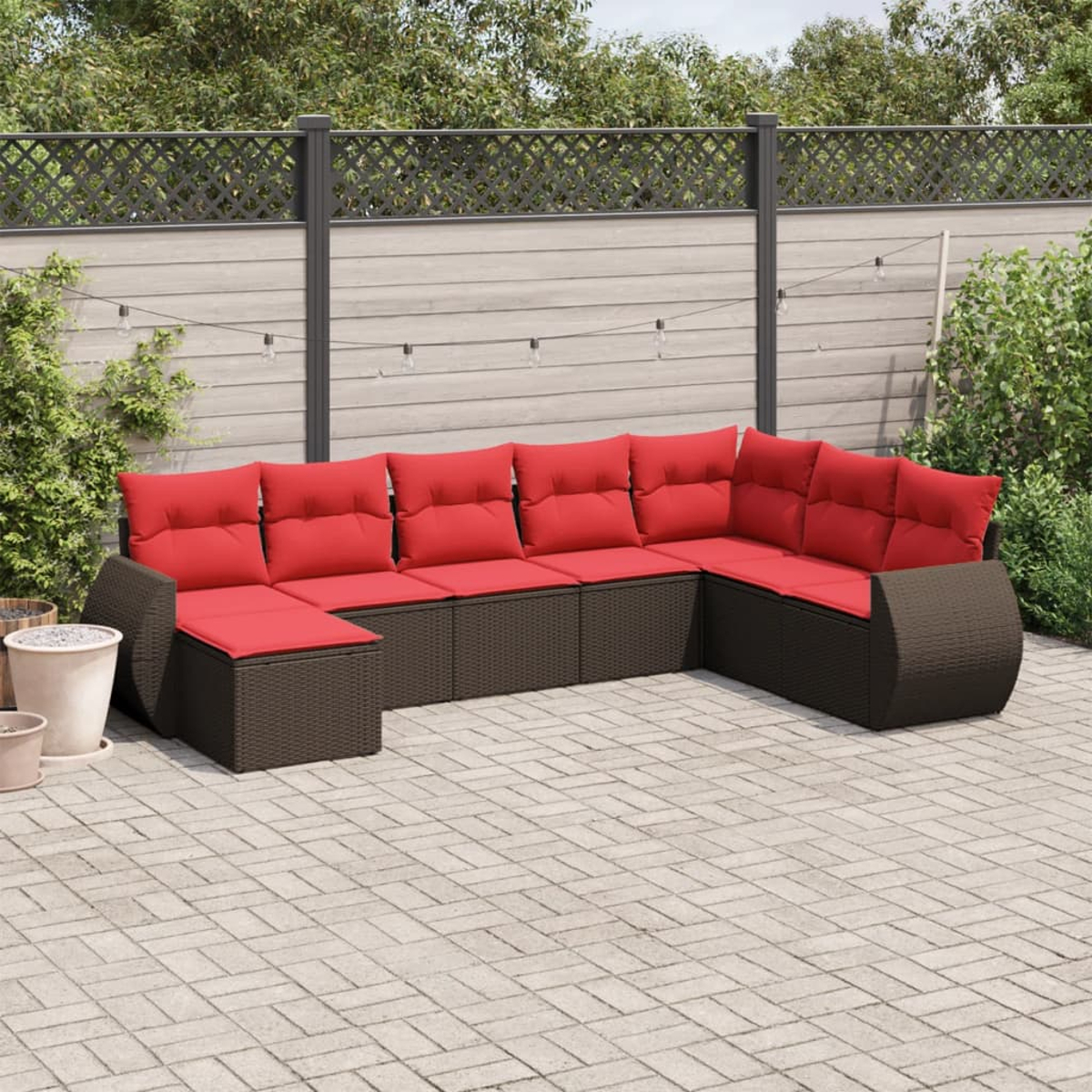 Ein braunes Korbsessel-Ecksofa mit roten Kissen steht auf einer Ziegelterrasse. Im Hintergrund befinden sich ein Zaun und Grün.