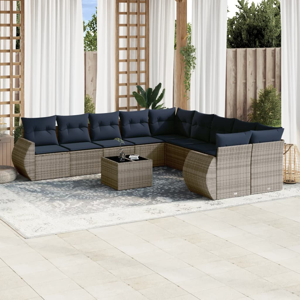 Außenbereich mit Ecksofa, Couchtisch und Teppich. Blaue Kissen kontrastieren mit den beigen Korbmöbeln. Vorhänge und Bäume sind im Hintergrund.