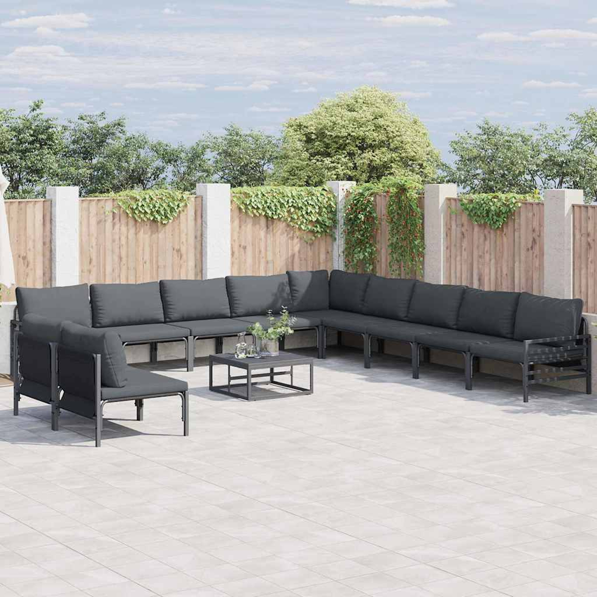 Outdoor-Ecksofa mit dunkelgrauen Kissen und einem niedrigen Tisch. Holzzaun und Laub im Hintergrund, auf einer Terrasse.