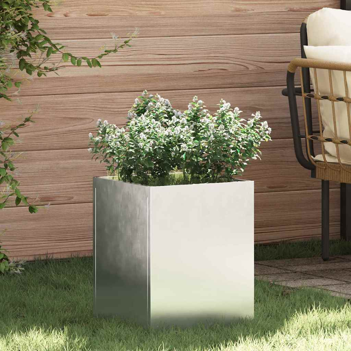 Ein quadratischer Metallpflanztopf mit grünen Pflanzen und weißen Blüten steht auf Gras. Eine Holzwand und ein Stuhl sind im Hintergrund.