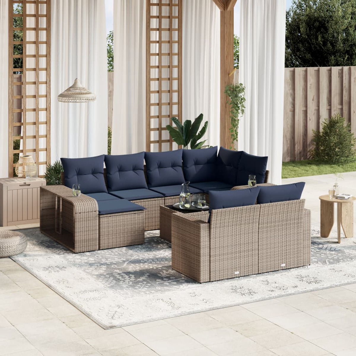 Outdoor-Sitzbereich mit gewebten Möbeln, blauen Kissen, einem gemusterten Teppich und transparenten Vorhängen. Ein Tisch hält Gläser und eine Wasserkaraffe.