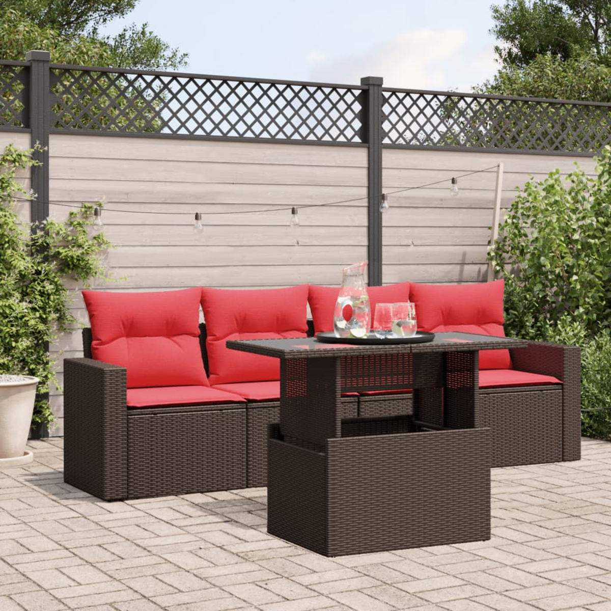 Garten-Set mit einem braunen, gewebten Tisch und Sofa mit roten Kissen. Auf dem Tisch stehen eine Kanne und Gläser. Zaun im Hintergrund.
