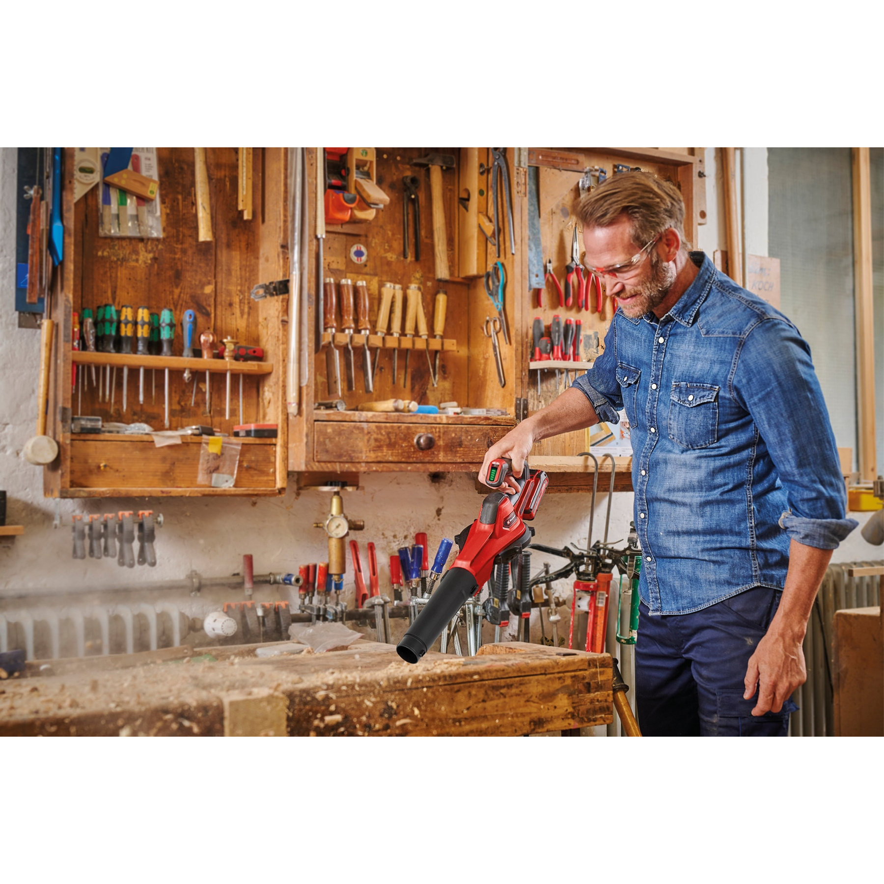 Mężczyzna w warsztacie używa czerwonej dmuchawy do liści na drewnianej powierzchni. Narzędzia na ścianie.