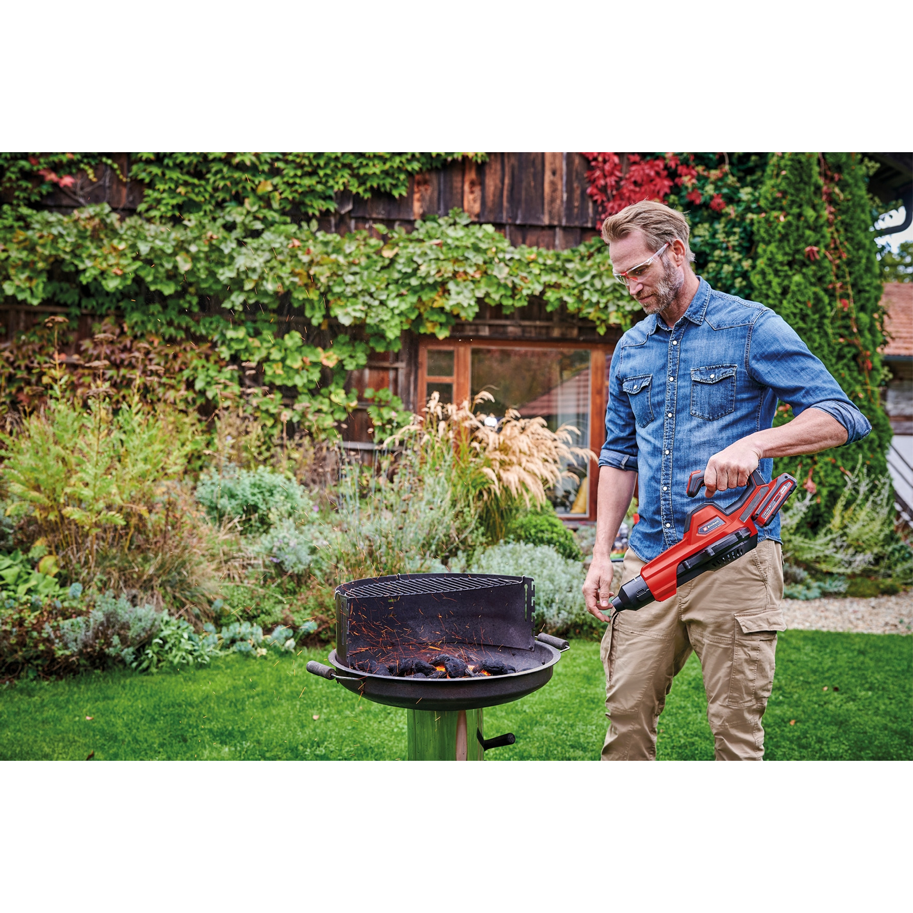 Mężczyzna używa czerwonej dmuchawy do grilla z płonącym węglem drzewnym. Ma okulary ochronne i stoi w ogrodzie.