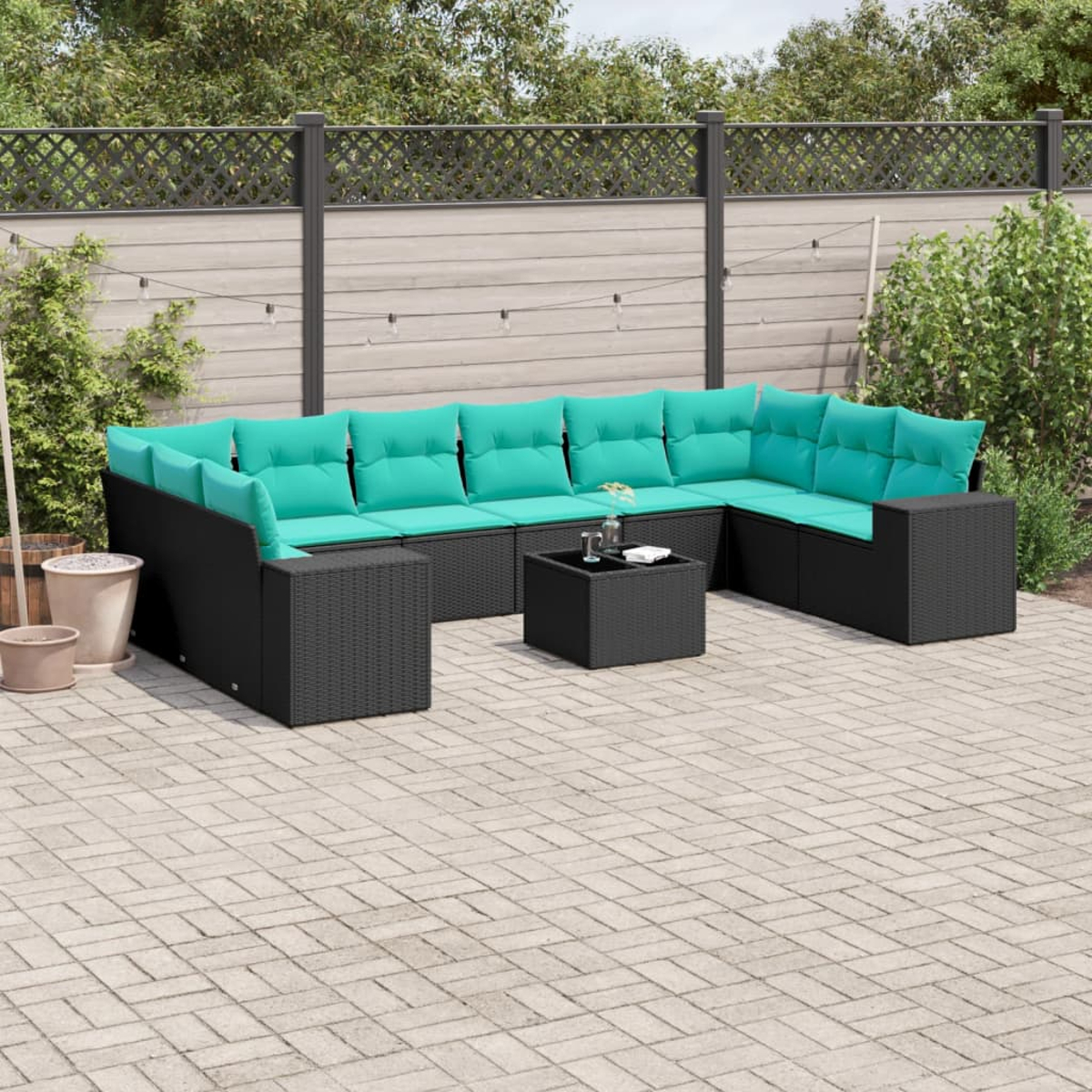 Schwarzes Outdoor-Ecksofa aus Korbgeflecht mit türkisfarbenen Kissen und kleinem Tisch auf einer Terrasse. Hintergrund: Zaun und Grün.